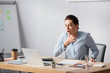 Brunette businesswoman with hand on neck coughing while sitting at workplace clipart