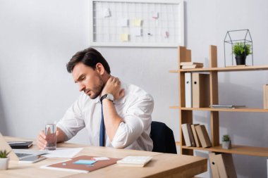 İşyerinde otururken, boynu ağrıyan bir iş adamı bir bardak su tutuyor.
