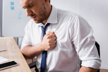 İş adamı kalp krizi geçirirken, iş yerinde bulanık arka planda oturuyor.
