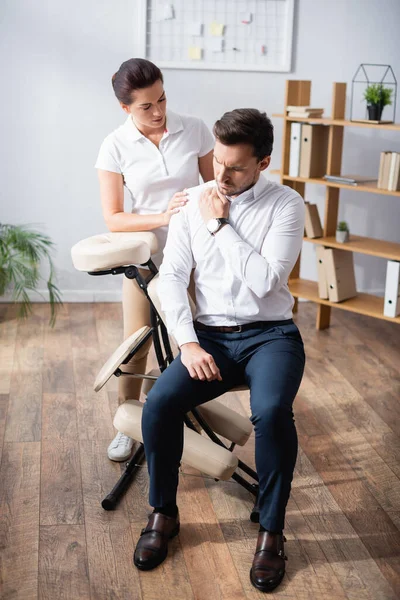 Massagista Olhando Para Ombro Doloroso Empresário Sentado Cadeira Massagem Escritório — Fotografia de Stock