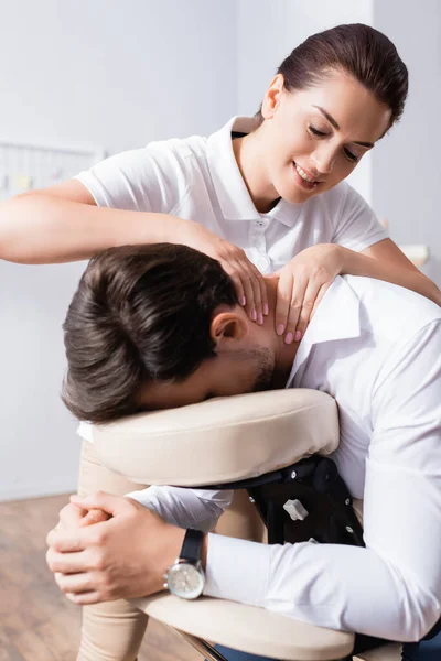 Smiling masseuse doing seated massage of businessman neck in office on blurred background