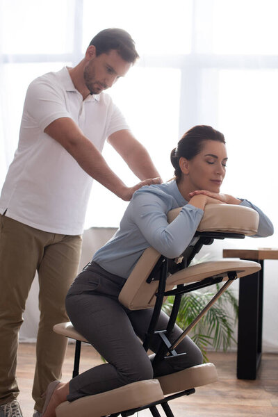 Masseur with outstretched hands massaging back of smiling woman with closed eyes sitting on massage chair in office