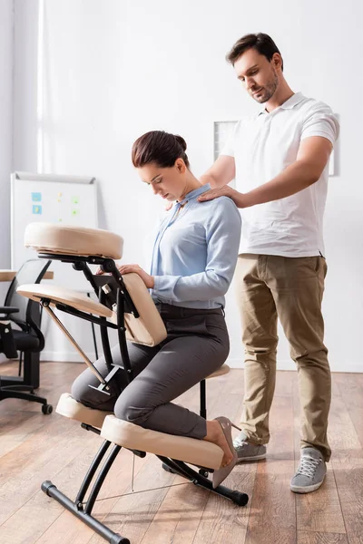 Massage therapist doing seated massage of shoulders for businesswoman sitting in office