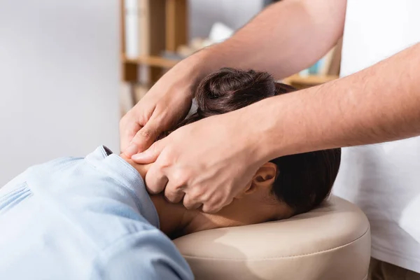 Cropped view of masseur doing seated massage of neck for brunette businesswoman in office on blurred background