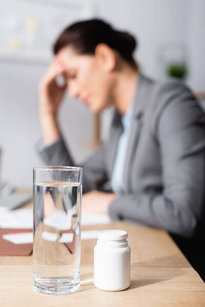 Close up view of glass of water and medication on desk with blurred businesswoman with headache on background