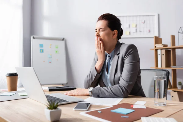 オフィスで職場に座っている間に口の近くに手を持つ眠そうなビジネスマンの女性 — ストック写真