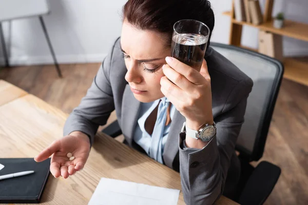 办公室女职员头疼 拿着一杯水 看着药丸 同时在模糊的背景下坐在工作场所 — 图库照片