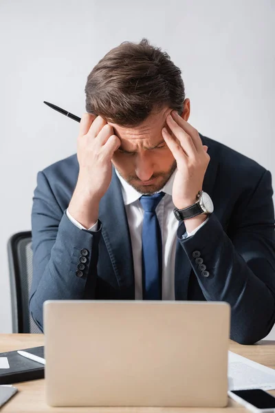 一位头疼的生意人手牵着手 坐在工作场所 面前放着模糊的笔记本电脑 — 图库照片