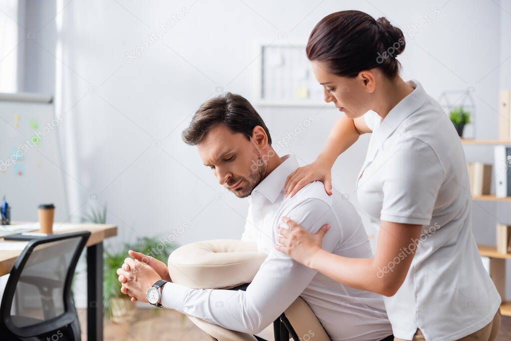 Female massage therapist massaging shoulder of client sitting on massage chair in office on blurred background