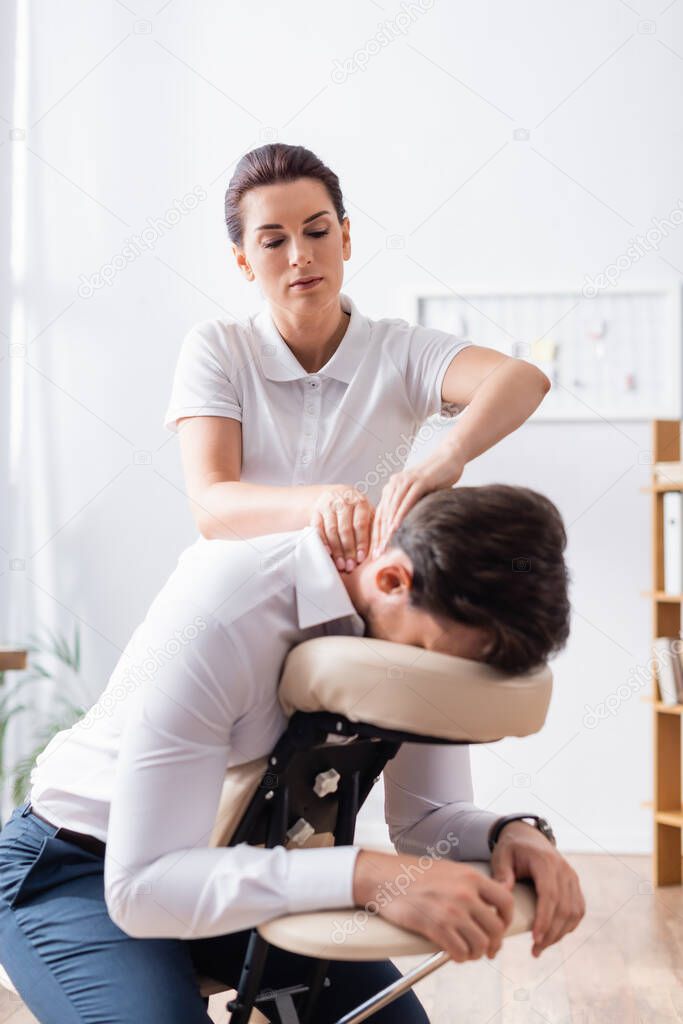 Female massage therapist massaging neck of businessman sitting on massage chair in office on blurred background