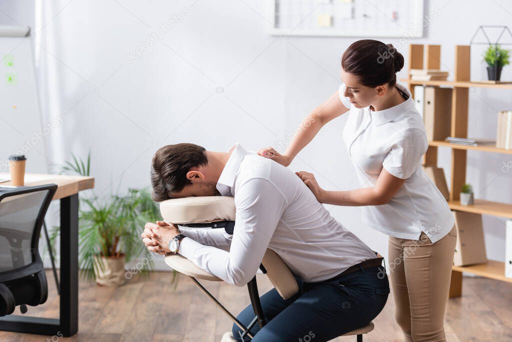 Side view of masseuse massaging back of businessman sitting on massage chair in office