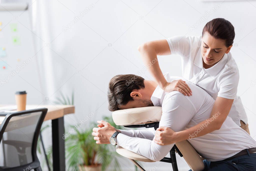 Positive masseuse doing arm massage for businessman in office on blurred background