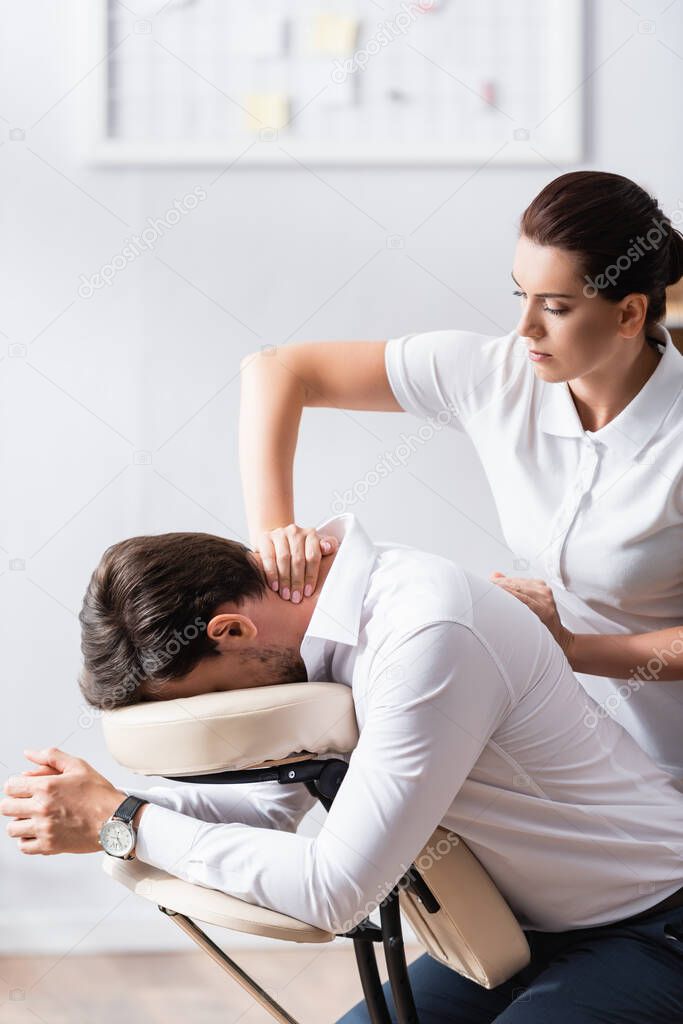 Masseuse massaging neck of businessman sitting on massage chair in office on blurred background
