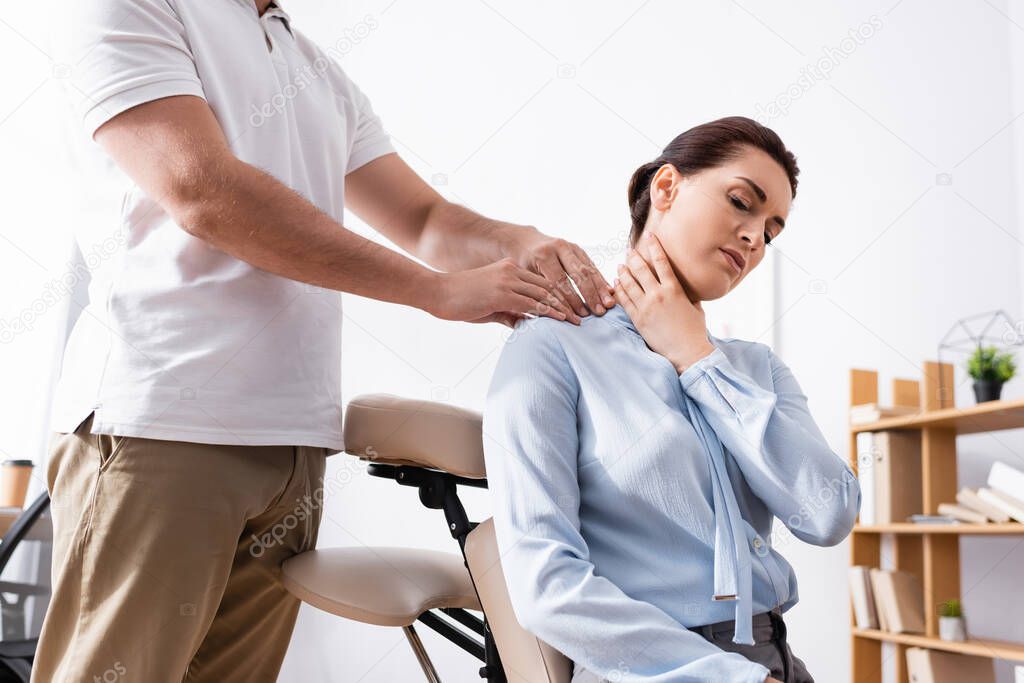 Massage therapist massaging painful shoulder of businesswoman siting on massage chair in office on blurred background