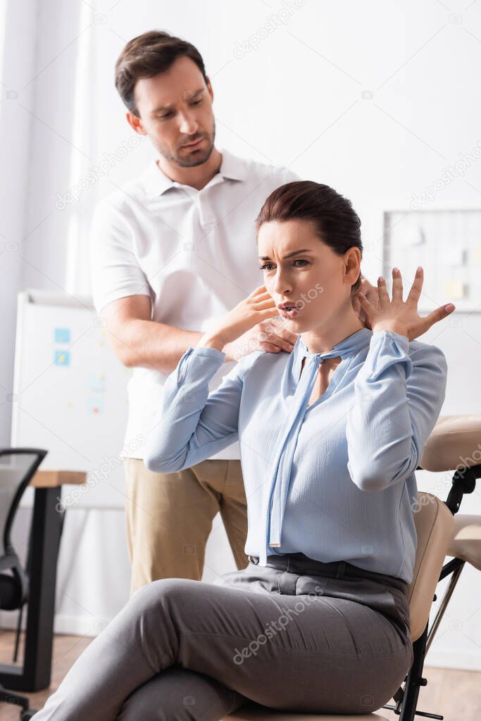 Masseur massaging painful neck of businesswoman complaining and gesturing with blurred office on background