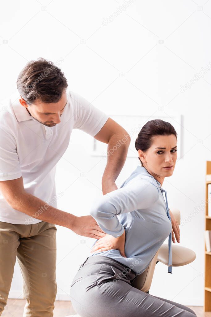 Masseur massaging painful back of businesswoman with hand on hip, sitting on massage chair on blurred background