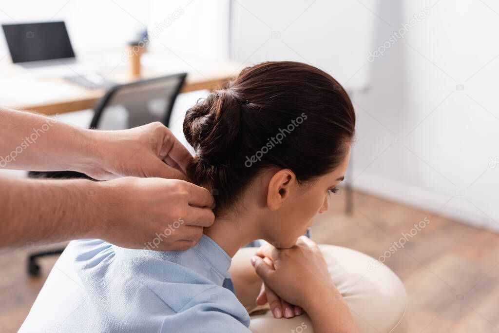 Masseur doing neck massage of brunette businesswoman sitting on massage chair on blurred background