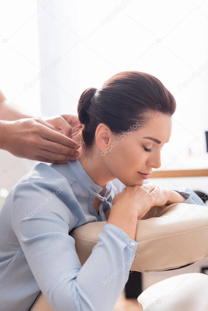 Masseur doing neck massage of female client with closed eyes on blurred background