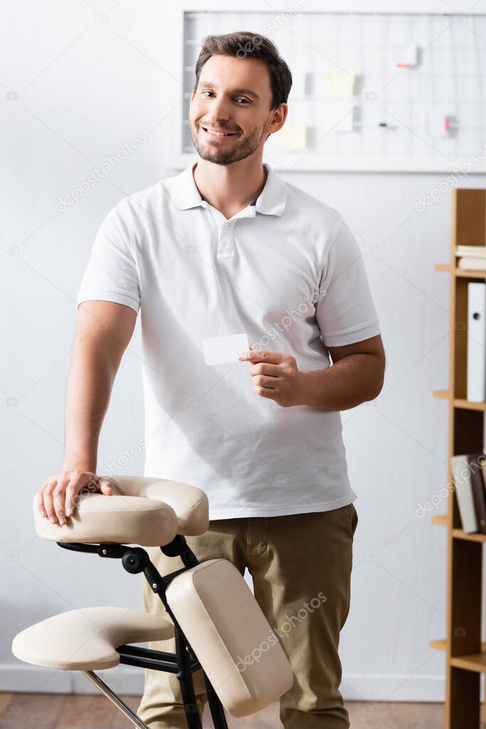 Smiling masseur showing blank business card while leaning on massage chair in office on blurred background