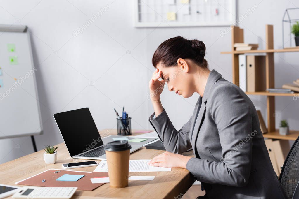 Tired businesswoman with hand on forehead sitting at desk near laptop and contract in office