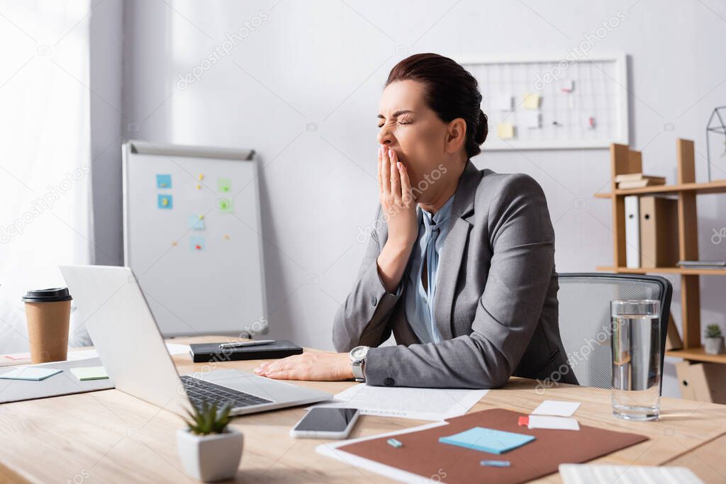 Sleepy businesswoman with hand near mouth yawning while sitting at workplace in office