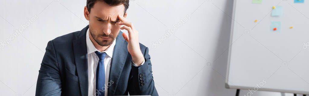 Tired businessman with headache, holding hand near temple and looking away with blurred flipchart on background, banner