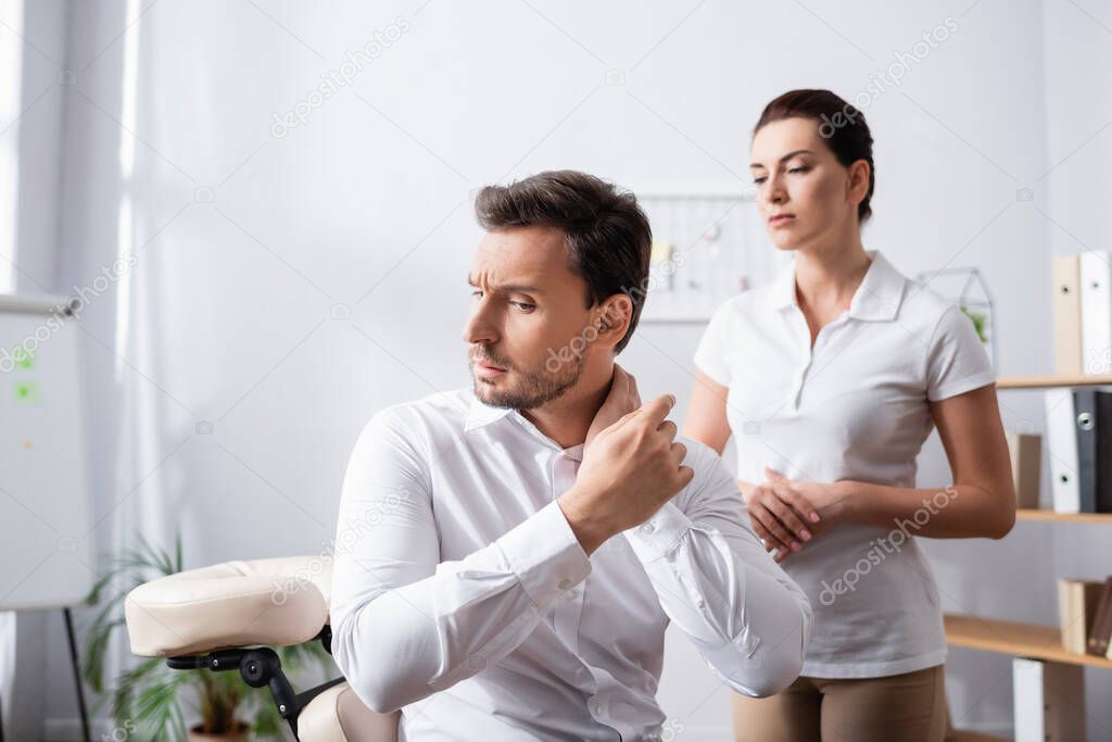 Masseuse looking at businessman pointing with finger at painful neck, while sitting on massage chair on blurred background