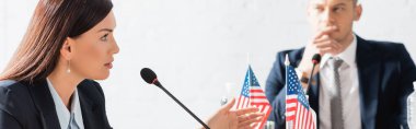 Kadın politikacı, mikrofonla konuşurken bulanık arka planda bulanık bir adamla Amerikan bayraklarının yanında oturuyor.