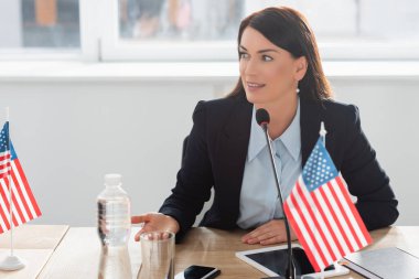 Smiling woman in formal wear looking away, while speaking in microphone, sitting near american flags in boardroom clipart