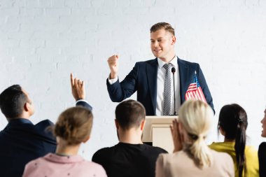 smiling political agitator showing win gesture in front of voters in conference hall clipart