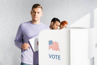man looking at camera while holding ballot near polling booth with american flag and vote lettering on blurred background clipart