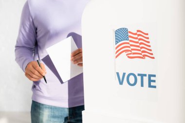 cropped view of man holding ballot and pen near polling booth with american flag and vote inscription clipart