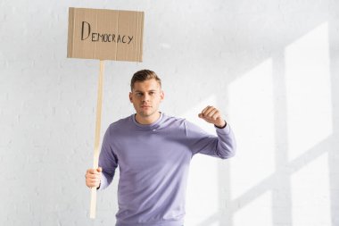 strict man with clenched fist holding placard with democracy inscription against white brick wall clipart