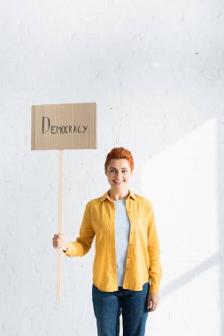 Beyaz tuğla duvara karşı demokrasi harfleriyle pankart tutan gülümseyen kadın.