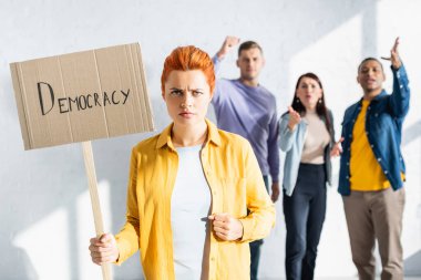 angry woman holding placard with democracy inscription while irritated multicultural people gesturing on blurred background clipart