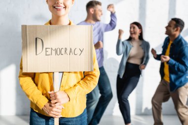 Elinde demokrasi yazısıyla gülümseyen bir kadın. Bulanık arka planda dans eden mutlu, çok kültürlü insanların yanında.