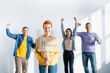 Elinde demokrasi yazılı pankart tutan gülümseyen bir kadın. Bulanık arka planda zafer jesti sergileyen çok kültürlü insanların yanında.