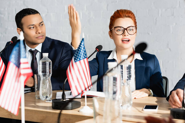 Indian politician looking at redhead woman with raised hand, while talking in boardroom on blurred foreground