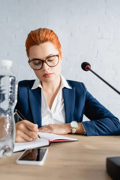 红头发女人穿着正式服装 写在笔记本上 坐在桌边 手里拿着一瓶水和话筒 — 图库照片