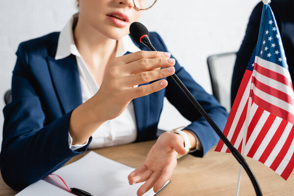 Обрезанный вид женщины-политика, говорящей в микрофоне, сидя за столом возле флага США в зале заседаний