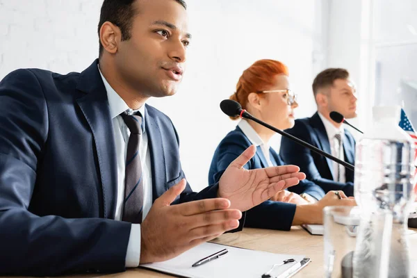 Indian Politician Gesturing While Speaking Microphone Boardroom Blurred Colleagues Foreground — Stock Photo, Image