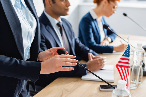 Женщина-политик касается микрофона, стоя рядом с коллегами в зале заседаний на размытом фоне