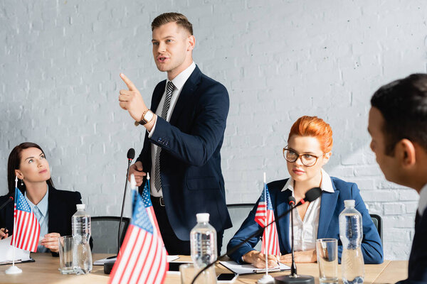 Smiling politician pointing with finger, speaking while standing near colleague writing on clipboard on blurred foreground