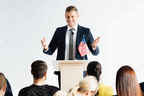smiling speaker standing with open arms in front of voters in conference hall, blurred foreground