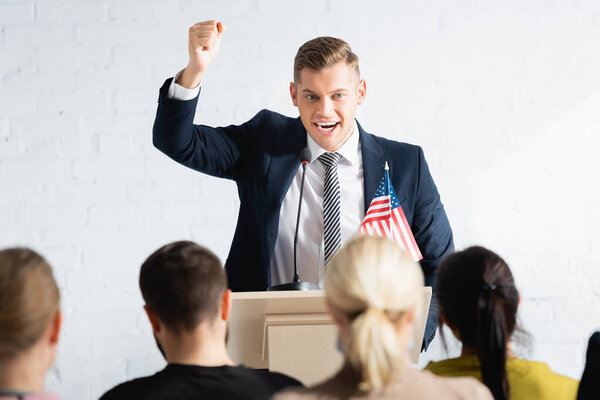 excited speaker showing winner gesture while talking to agitators in conference hall, blurred foreground