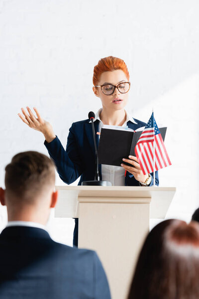 speaker looking at notebook and gesturing in front of voters in conference room, blurred foreground