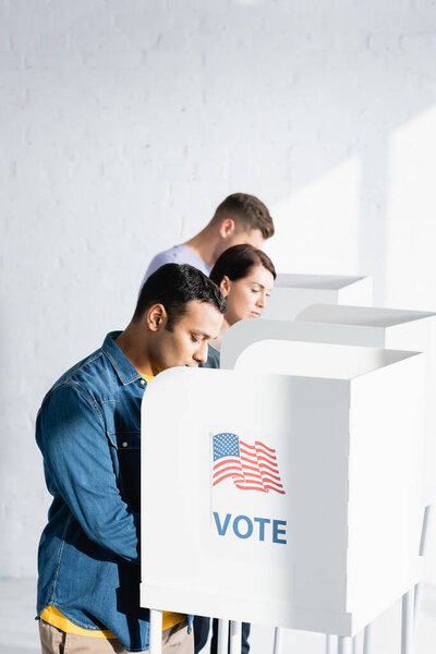 multicultural voters in polling cabins with american flag and vote inscription on blurred background