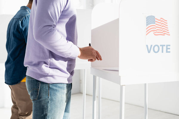 cropped view of multicultural men in polling booth with american flag and vote inscription 