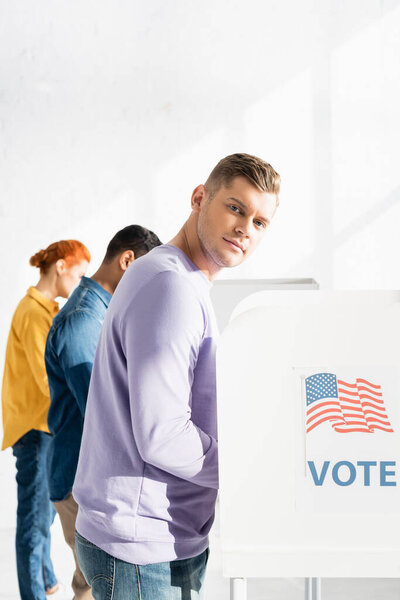 man looking at camera near polling booth with american flag and vote inscription, and multicultural electors on blurred background