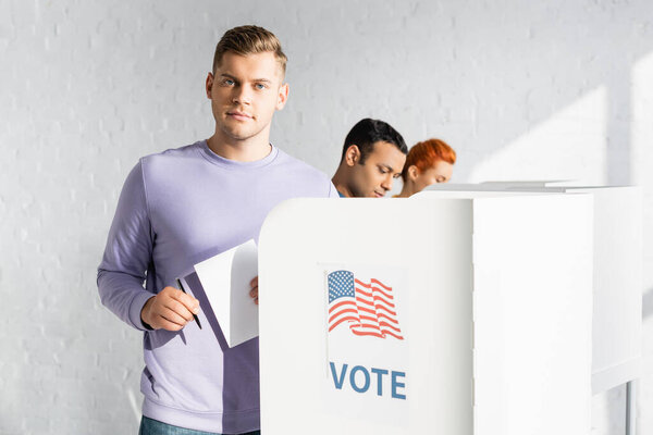 man looking at camera while holding ballot near polling booth with american flag and vote lettering on blurred background